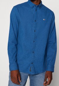 Blaues Hemd mit Knopfleiste aus strukturiertem Material und Kragen, mit schwarzen Knöpfen und einem kleinen Logo auf der Brust. Lange Ärmel.