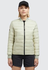 Light green padded jacket with a bomber style, black zipper, and side pockets; features ribbed cuffs and a logo on the chest. Black cap worn.