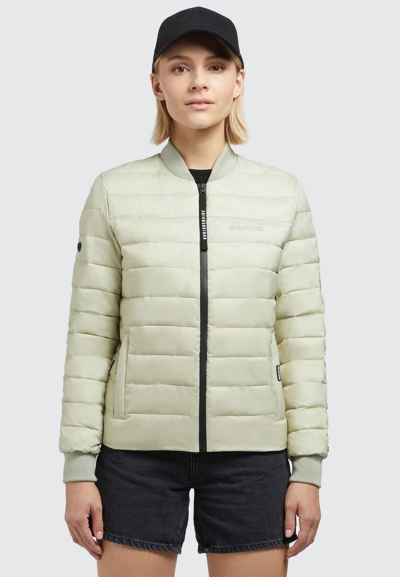 Light green padded jacket with a bomber style, black zipper, and side pockets; features ribbed cuffs and a logo on the chest. Black cap worn.