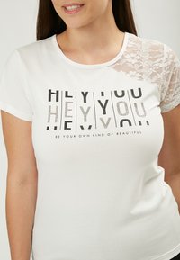 Witte t-shirt met korte kanten mouwen, met een grafisch ontwerp in zwarte tekst met de tekst "HEY YOU" en de frase "WEES JE EIGEN SOORT VAN MOOI."