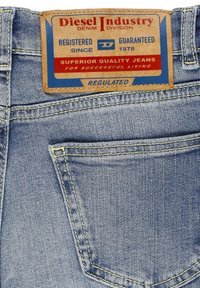 Jeans di denim azzurro chiaro con una toppa in pelle marrone decorata con grafiche rosse e blu. Cuciture a contrasto e un piccolo dettaglio di tasca posteriore visibile.