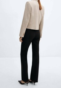 Blazer beige accorciato con spalle strutturate e dettagli in bottone dorato, abbinato a pantaloni neri slim-fit e décolleté neri a punta.