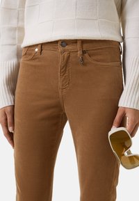 Braune Cordhosen mit schmaler Passform, ausgestattet mit frontalen Taschen und einem Reißverschluss. Eine weiße Hand hält übergroße Sonnenbrillen in der Nähe der Taille.