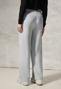 Pantalons large gris clair avec une texture lisse, une taille élastique et deux poches arrière, associés à des chaussures légères sur un fond neutre.