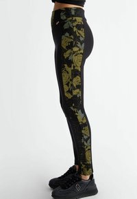 Leggings negros con un patrón floral verde en el lateral, elaborados con una tela elástica. Combinados con zapatillas negras texturizadas.