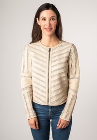 Giacca in pelle beige con motivo a strisce diagonali e accenti argentati, con scollo rotondo e chiusura con zip, abbinata a jeans blu.