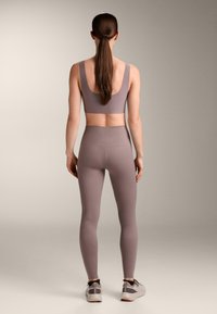 Ženska z dolgimi rjavimi lasmi v nizkem čopku, oblečena v vijoličast športni modrček, visokopasne legice in svetlo sive tekaške copate, stoji z obrazom stran.