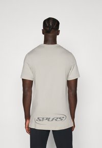 Camiseta de color gris claro con mangas cortas, cuello redondo y dobladillo curvado. Presenta un logo en color gris oscuro que dice "SPURS" impreso en la parte posterior.
