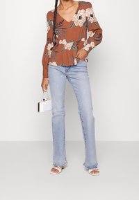 Žena v rustikální květinové blůze, světle modrých rovně střižených džínách, bílé sandálech na podpatku a držící malou bílou kabelku.