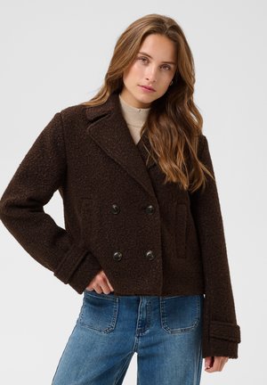 Femme portant une veste en laine texturée brun foncé à double boutonnage et un jean bleu, posant avec une main sur la hanche.