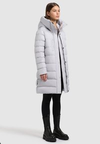 Šviesiai pilka pūkinė striukė su aukštu apykakle, horizontaliu dygsniu, priekyje užsegama mygtukais ir šoninėmis kišenėmis. Dėvima ant juodų leginsų.