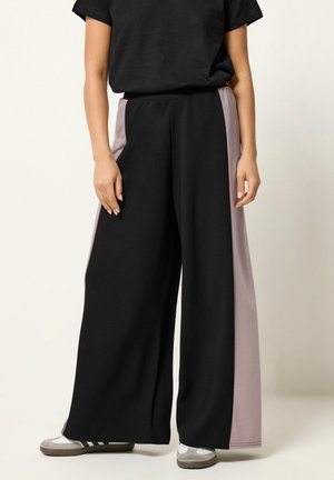 Osoba nosząca czarne spodnie o szerokich nogawkach z szarymi panelami bocznymi, czarną bluzkę z krótkim rękawem, białe skarpetki i szare trampki, stojąca na jednolitym tle.