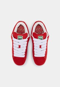 Red suede sneakers with white laces, featuring a padded tongue and collar. Green logo tag on the tongue, patterned insoles. Classic design.