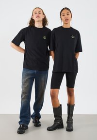 Sorte oversized t-shirts med grønt logo, lyseblå slidte jeans, sorte leggings og knælange sorte støvler med dekorativ hardware.