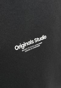 Tessuto di cotone nero con testo bianco: "Originals Studio," "Portato a te da JACK&JONES," e "Progettato a Copenaghen." Texture liscia.