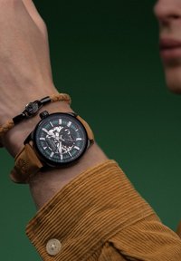 Montre analogique noire avec un cadran transparent, des accents verts et argentés, et un bracelet en cuir marron ; portée avec un bracelet tressé.