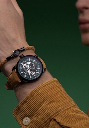 Montre analogique noire avec un cadran transparent, des accents verts et argentés, et un bracelet en cuir marron ; portée avec un bracelet tressé.
