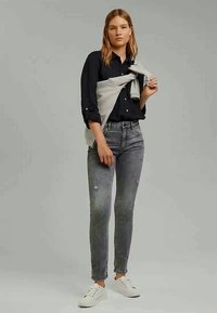 Camisa preta de botões, suéter cinza claro amarrado à cintura, jeans skinny cinza desgastados e ténis brancos.