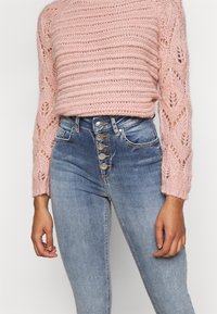 Suéter de malha rosa claro com mangas texturizadas, combinado com jeans de ganga azul de cintura alta com fecho de botão à frente.