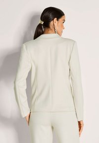 Vrouw met donker haar in een paardenstaart, gekleed in een crème kleurig op maat gemaakt blazer en bijpassende broek, staande tegen een effen lichte achtergrond.