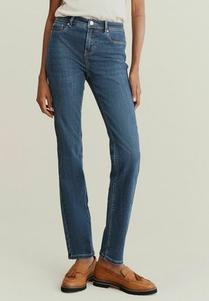 Vrouw die een mid-rise rechte blauwe jeans en beige loafers met kwastjes draagt, staand tegen een effen lichte achtergrond.