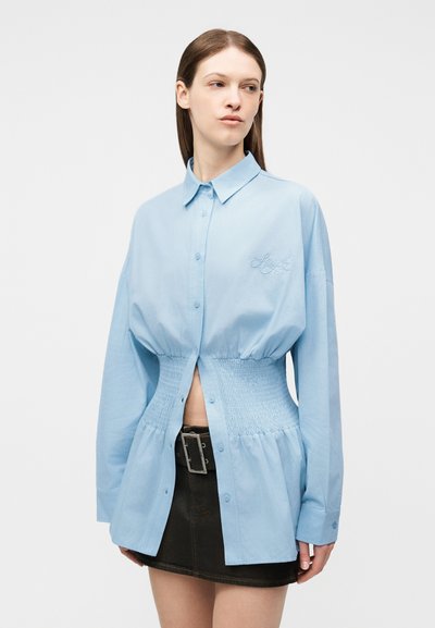 Γυναίκα που φοράει μπλε ανοιχτό πουκάμισο με κουμπιά και λεπτομέρεια με ελαστική μέση, καθώς και μαύρη μίνι φούστα με φαρδιά ζώνη και αγκράφα.