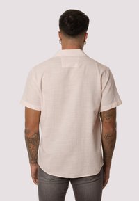 Camisa de manga corta en tejido rosa claro, de textura ligera, con canesú en la parte posterior y un corte relajado. Presenta mangas dobladas y un dobladillo curvado.