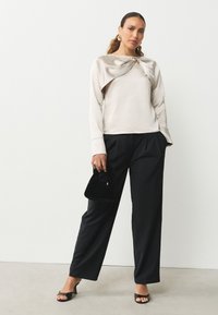 Top beige claro con detalle frontal retorcido, combinado con pantalones anchos negros. La modelo sostiene un pequeño bolso de terciopelo negro y lleva sandalias de tacón.