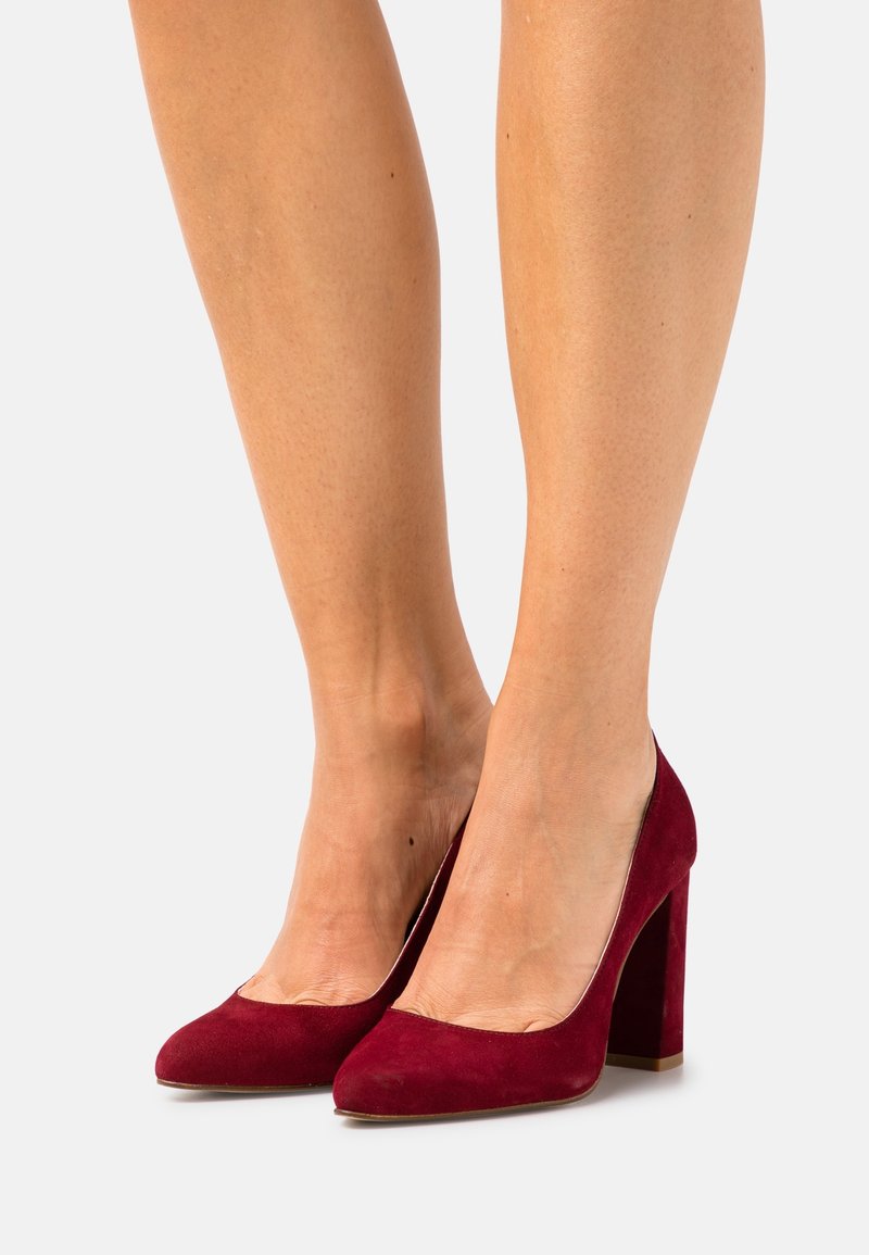 Escarpins en daim bourgogne à talons hauts avec un bout rond et un talon épais. La texture est douce, et les chaussures ont un design élégant et simple.