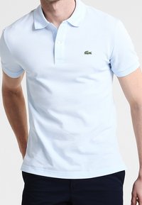 Helles blaues Kurzarm-Poloshirt aus Baumwolle mit einer Dreiknopf-Leiste und kleinem grünen gestickten Logo auf der linken Brust.