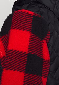 Czarna pikowana kurtka z czerwono-czarną kraciastą naszywką z polaru na lewym rękawie, łącząca kontrastujące faktury i wzory.