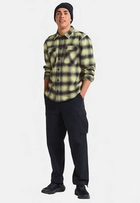 Camicia di flanella a quadri gialla e nera, con due tasche sul petto, abbinata a pantaloni cargo neri. Il modello indossa sneakers nere e un berretto.