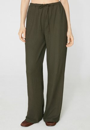 Mujer con pantalones anchos de textura verde oscuro y cintura con cordón, y zapatos marrones, de pie frente a un fondo liso.