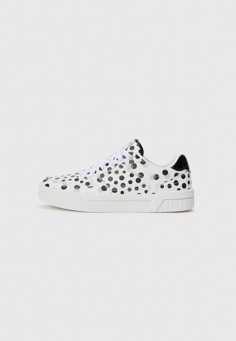 Sneaker basse blanche avec des pois noirs, lacets blancs, semelle épaisse et languette de talon noire brillante sur un fond uni.