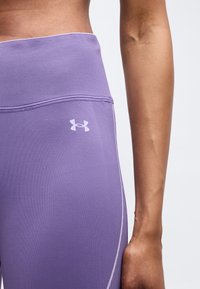 Κοντινό πλάνο ενός ατόμου που φοράει μωβ κολάν Under Armour με ψηλή μέση και λογότυπο στο αριστερό ισχύ, με το χέρι ορατό δίπλα στο σώμα.