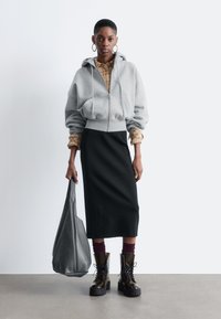 Grau zip-up Kapuzenpullover, kariertes Hemd darunter, schwarzer Midirock, klobige schwarze Stiefel, hält eine große graue Ledertasche. Überall glatte Texturen.