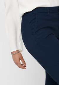 Pantalons extensibles bleu marine avec une coupe slim, poches latérales et détails de couture visibles, assortis à un haut à manches longues blanc.