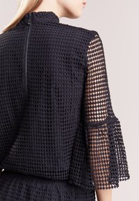 Top negro con un patrón de crochet texturizado, elevado cuello, y mangas acampanadas transparentes con un borde en forma de concha. Incluye un cierre de cremallera en la parte posterior.