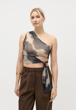 Frau mit blondem Haar trägt ein ein-ärmeliges Batik-Crop-Top und hoch taillierte braune Hose, accessoriert mit goldenen Armband und Ohrringen.