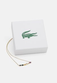 Gouden ketting met kleurrijke geometrische edelstenen en bedel, gepresenteerd bovenop een gestructureerde witte doos met een groen krokodillenlogo.