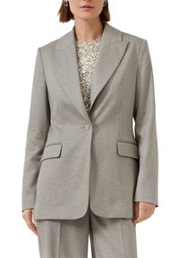 Vrouw in een op maat gemaakt lichtgrijs blazer met klepzakken en een enkele decoratieve knoop, gecombineerd met bijpassende broeken en een top met pailletten.