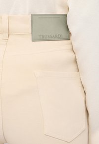 Pantalones de mezclilla beige con un bolsillo trasero visible y una etiqueta verde que dice "TRUSSARDI". La tela muestra una textura fina y un acabado suave.