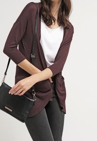 Žena v tmavě červeném kardiganu přes bílý top, držící černou koženou crossbody tašku na jednobarevném pozadí.
