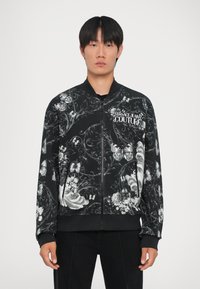 Giacca bomber nera con intricati motivi floreali e farfalli bianchi, polsini e colletto a coste, chiusura frontale con zip e patch con logo.