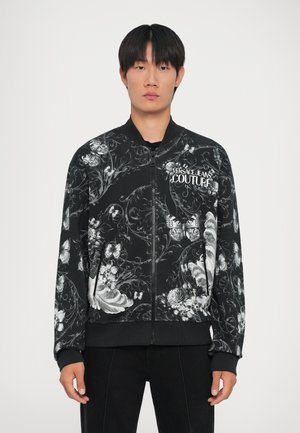 Schwarze Bomberjacke mit aufwendigen weißen Blumen- und Schmetterlingsmustern, gerippten Bündchen und Kragen, vorne mit Reißverschluss und Logopatch.