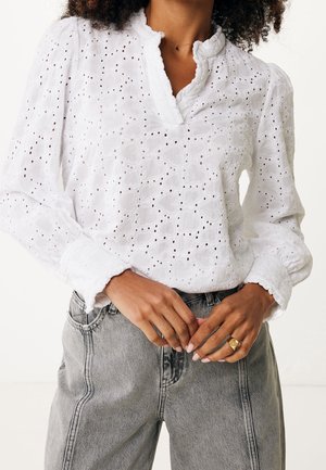 Femme portant un chemisier blanc en dentelle œillet avec un col et des poignets à volants, associé à un jean gris taille haute, les mains jointes montrant une bague en or.