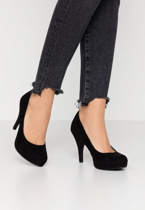 Stopy w czarnych zamszowych szpilkach w połączeniu z ciemnoszarymi skinny jeansami z postrzepionymi brzegami na białej podłodze.