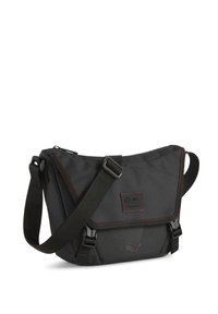 Sac bandoulière noir en matériau durable, avec des coutures rouges, une sangle réglable, un fermoir rabat et un patch logo.