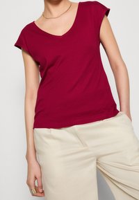 Burgundy T-shirt with short sleeves and a V-neck design, made from soft fabric, paired with light beige trousers. Smooth texture.