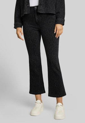 Frau trägt schwarze kurze Schlagjeans mit dezentem Leopardenmuster, weiße Sneaker und eine passende schwarze Jacke über einem weißen Top.